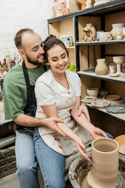 Sorridente ragazza artigianale abbracciando mentre fa vaso di argilla sulla ruota in ceramica in studio di ceramica — Foto stock