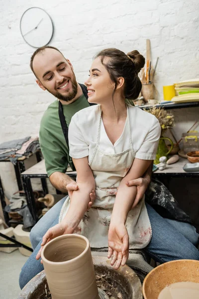 Positiva novia abrazo artesanal en delantal y hacer jarrón de arcilla en la rueda de cerámica en el estudio - foto de stock