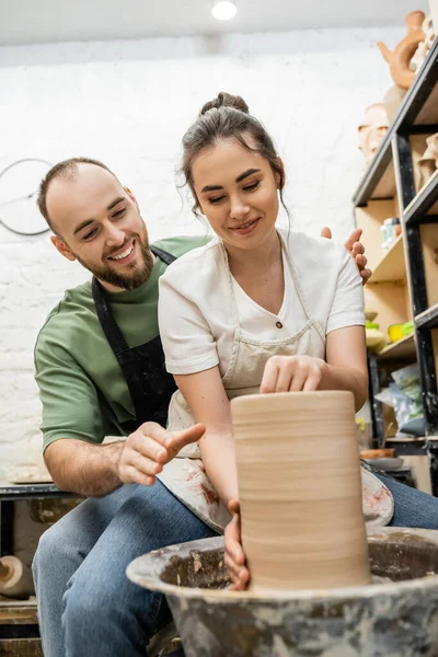 Artesano sonriente en delantal hablando con su novia haciendo jarrón de barro en rueda de cerámica en el taller - foto de stock