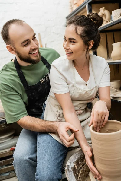 Artesanos sonrientes mirándose unos a otros y haciendo jarrón de barro sobre rueda de cerámica en estudio de cerámica - foto de stock