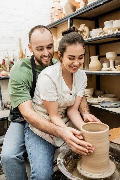 Alegre artesano abrazando novia y haciendo jarrón de arcilla juntos en la rueda de cerámica en el taller - foto de stock