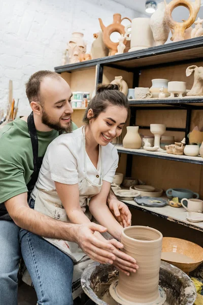 Craftsman in apron shaping clay vase on pottery wheel with smiling girlfriend in ceramic workshop — Stock Photo