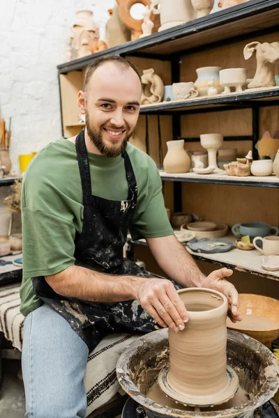 Улыбающийся бородатый ремесленник в фартуке смотрит в камеру и формирует глиняную вазу на гончарном круге в студии — стоковое фото