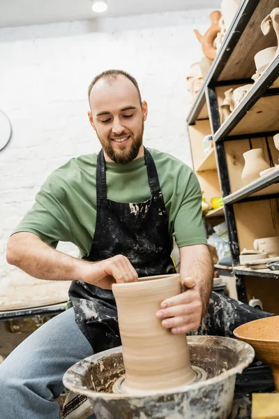 Smiling barbudo artesano masculino en delantal que forma la escultura de arcilla en la rueda de cerámica en taller de cerámica - foto de stock