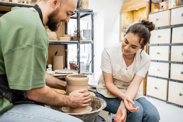 Улыбающаяся ремесленница в фартуке сидит рядом с парнем и делает глиняную вазу на гончарном круге в мастерской — стоковое фото