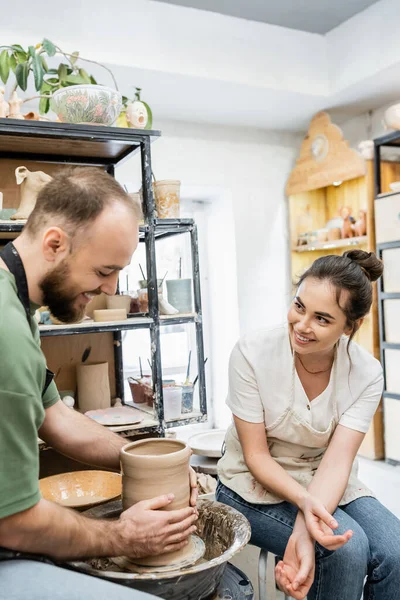 Joyful artisan in apron talking to boyfriend making clay vase on pottery wheel in ceramic workshop — Stock Photo