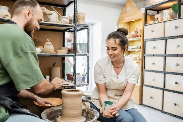 Artesana sonriente sosteniendo la taza mientras su novio hace jarrón de arcilla en la rueda de cerámica en el estudio - foto de stock