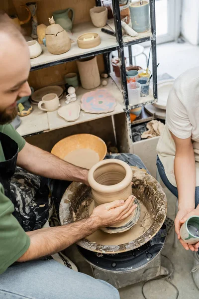 Vista de alto ângulo do vaso de barro de modelagem artesanal na roda de cerâmica perto da namorada com copo no estúdio — Fotografia de Stock