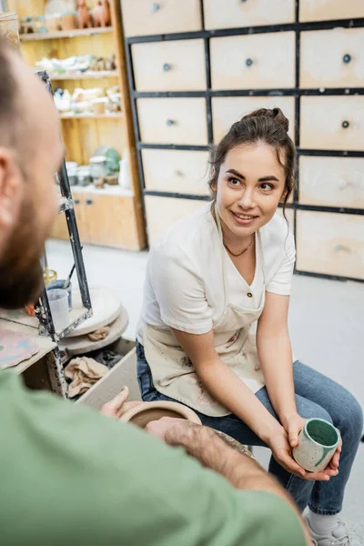 Artesã sorridente em avental segurando copo perto namorado borrado fazendo vaso de barro na roda de cerâmica — Fotografia de Stock