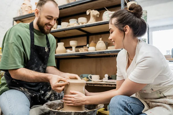 Smiling romantic artisans in aprons shaping clay vase together on pottery wheel in ceramic workshop — Stock Photo