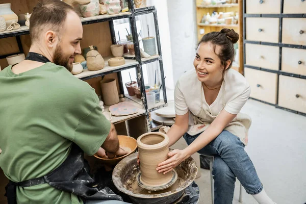 Sourire artisan façonnant vase d'argile sur la roue de poterie près du petit ami et bol avec de l'eau en studio — Photo de stock