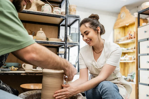 Радостная ремесленница в фартуке делает глиняную вазу с размытым парнем на гончарном круге в мастерской — стоковое фото