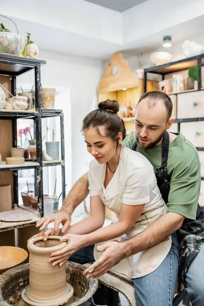 Artigiano barbuto che parla con gioiosa fidanzata plasmando vaso di argilla sulla ruota in ceramica in studio di ceramica — Foto stock