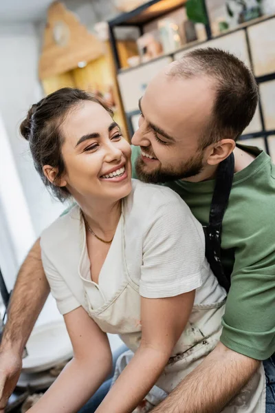 Artesãos sorridentes em aventais abraçando e olhando uns para os outros em oficina de cerâmica borrada — Fotografia de Stock