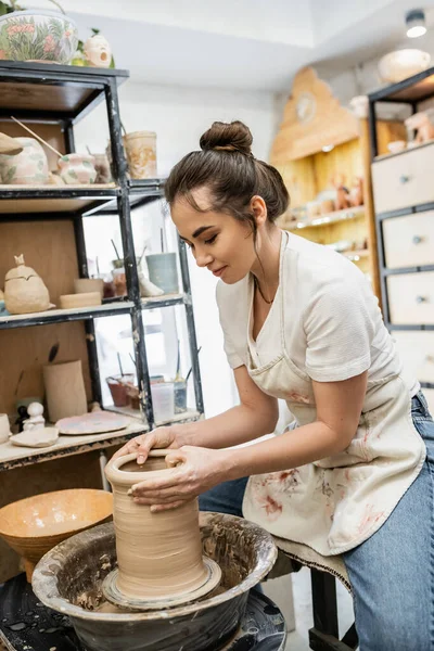 Ceramista bruna in grembiule che crea vaso di argilla sulla ruota della ceramica vicino alla ciotola con acqua in officina — Foto stock
