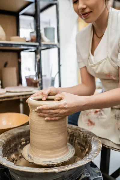 Vista cortada de oleiro fêmea em avental criando vaso de barro na roda de cerâmica em oficina turva — Fotografia de Stock