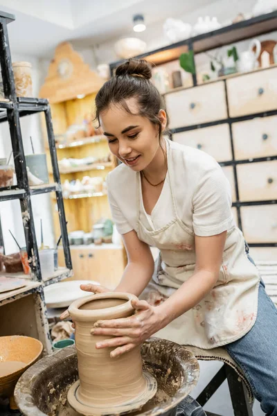 Artisanat féminin positif en tablier façonnant vase d'argile sur roue de poterie en atelier céramique en arrière-plan — Photo de stock