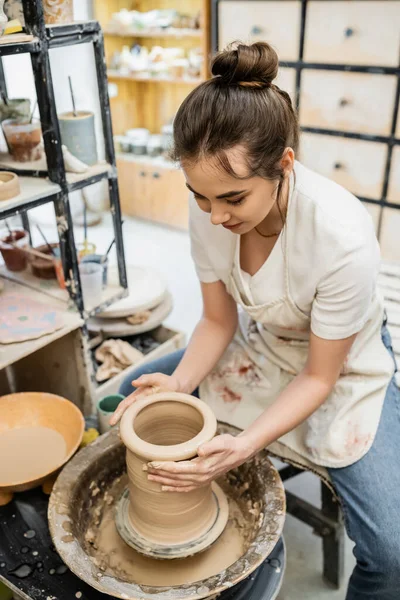 Brünette Kunsthandwerkerin in verschwommener Schürze macht Tonvase auf Töpferscheibe in Wassernähe in Werkstatt — Stockfoto