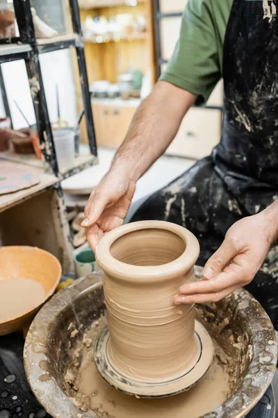 Vista recortada del artesano masculino en delantal formando jarrón de arcilla sobre rueda de cerámica cerca del agua en taller - foto de stock