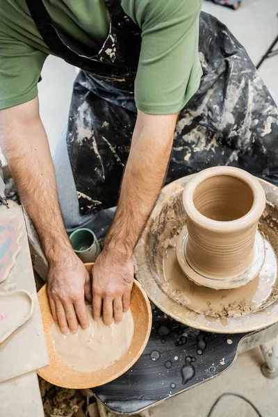 Vue du dessus du potier masculin dans le tablier travaillant avec de l'eau dans un bol et de l'argile sur une roue de poterie en atelier — Photo de stock