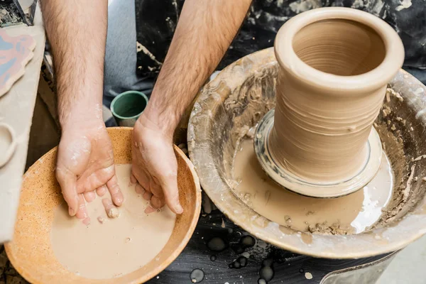 Vue du dessus de l'artisan en tablier travaillant avec de l'eau dans un bol et de l'argile sur une roue de poterie en studio — Photo de stock