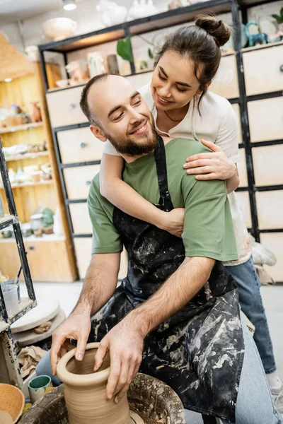 Artigiana sorridente in grembiule che abbraccia fidanzato plasmando argilla sulla ruota in ceramica in studio di ceramica — Foto stock