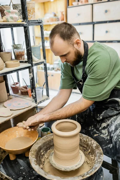 Бородатый скульптор в фартуке моет руки в миске с водой возле глины и гончарного колеса в мастерской — стоковое фото