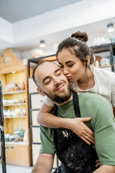Feminino artesanal beijando e abraçando namorado em avental enquanto trabalham juntos em oficina de cerâmica — Fotografia de Stock