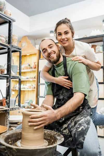 Smiling craftswoman hugging boyfriend shaping clay on pottery wheel and looking at camera in studio — Stock Photo