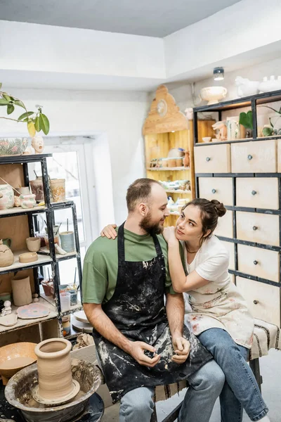 Brunette craftswoman in apron hugging boyfriend  near clay on pottery wheel in ceramic workshop — Stock Photo