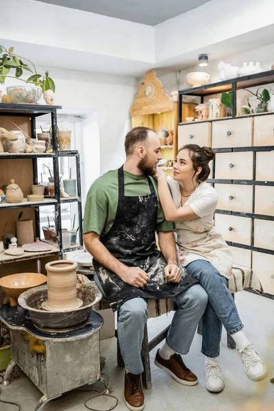 Craftswoman in apron hugging boyfriend and talking near clay on pottery wheel in workshop — Stock Photo