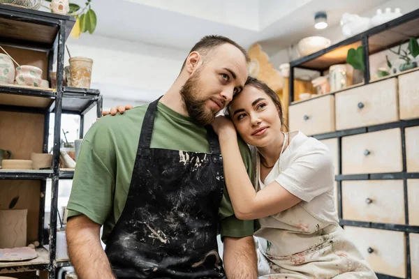 Craftswoman no avental abraçando namorado barbudo e olhando para longe no estúdio de cerâmica no fundo — Fotografia de Stock