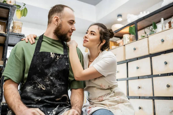 Craftswoman hugging and looking at boyfriend in apron while working in ceramic workshop — Stock Photo