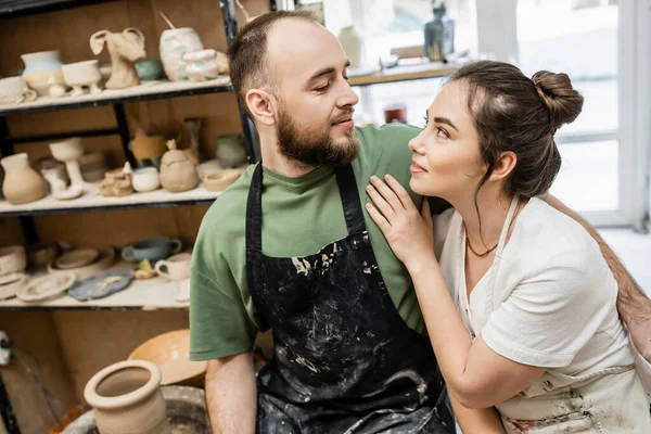 Bearded craftsman in apron hugging and looking at girlfriend near blurred clay sculptures in studio — Stock Photo