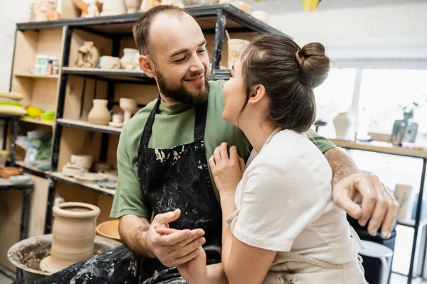 Улыбающийся ремесленник в фартуке обнимает и держит за руку улыбающуюся девушку в размытой керамической мастерской — стоковое фото