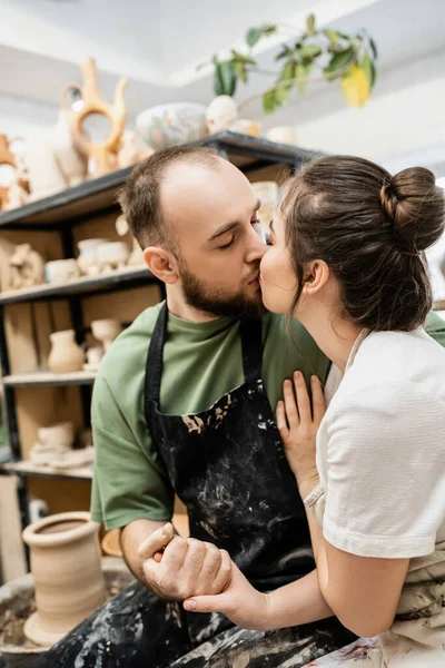 Artesanato no avental beijando e segurando a mão da namorada perto da roda de cerâmica borrada na oficina — Fotografia de Stock