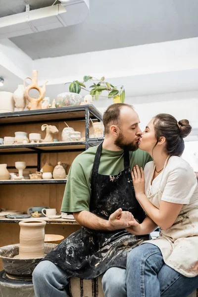 Casal de escultores em aventais de mãos dadas e beijando perto da roda de cerâmica na oficina — Fotografia de Stock