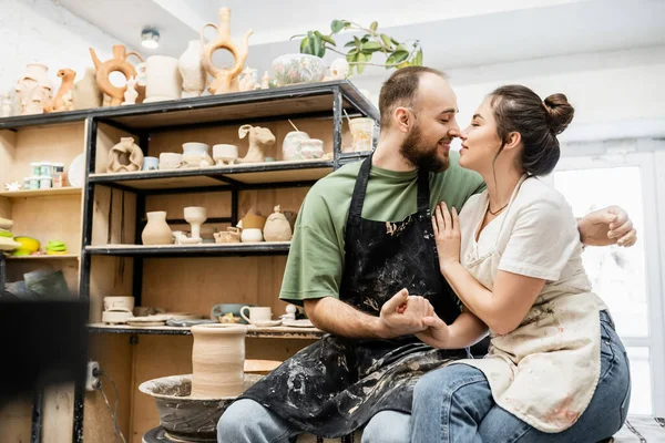 Vista lateral do casal de artesãos em aventais de mãos dadas e sorrindo perto da roda de cerâmica no estúdio — Fotografia de Stock