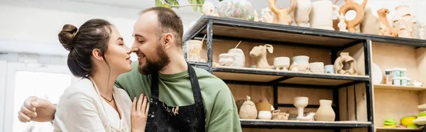 Artisanat souriant dans tablier embrasser petite amie tout en travaillant ensemble dans un atelier de céramique, bannière — Photo de stock