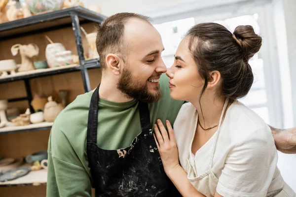 Vista lateral do escultor sorridente no avental beijando namorada morena na oficina de cerâmica borrada — Fotografia de Stock