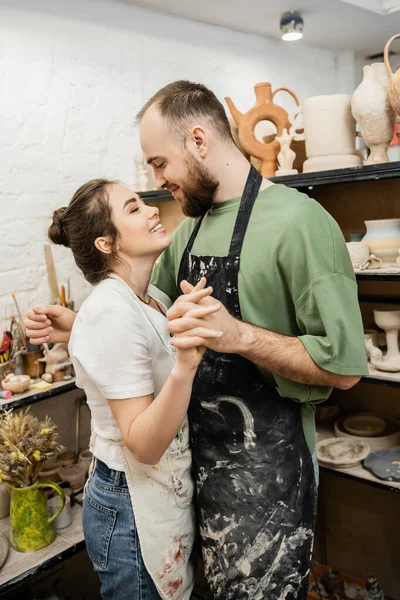 Smiling couple of artisans in aprons holding hands and hugging in ceramic workshop — Stock Photo