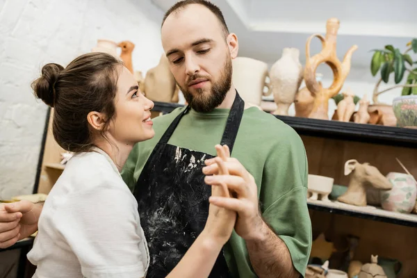 Бородатый скульптор в фартуке держит за руку улыбающуюся девушку во время работы в керамической мастерской — стоковое фото