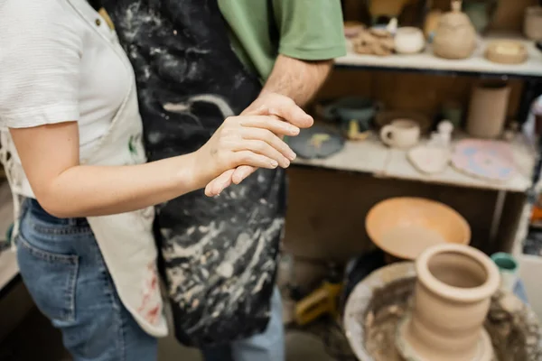 Vista cortada de casal de escultores em aventais de mãos dadas perto da roda de cerâmica borrada na oficina — Fotografia de Stock