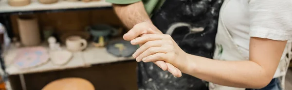 Vista recortada de un par de alfareros en delantales tomados de la mano mientras trabajan en taller de cerámica, pancarta - foto de stock