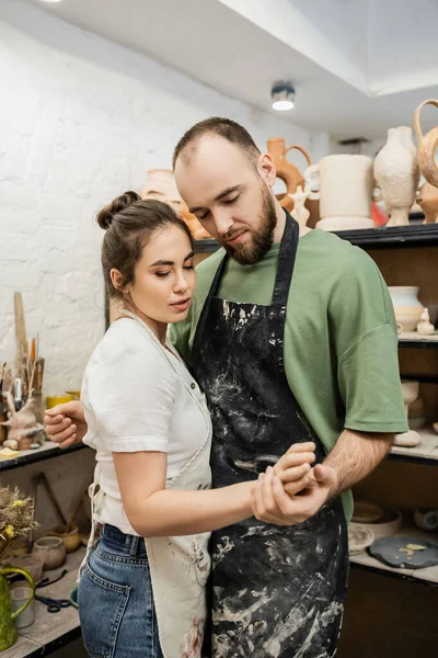 Couple de sculpteurs dans des tabliers tenant la main tout en travaillant ensemble dans un atelier de poterie — Photo de stock