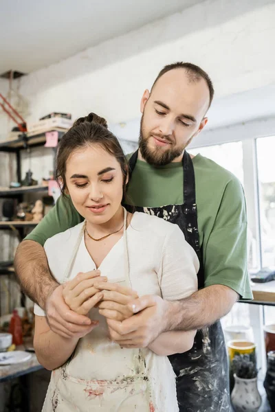 Бородатый мастер, держащийся за руки и обнимающий улыбающуюся девушку в фартуке в керамической мастерской — стоковое фото