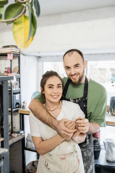 Lächelndes Bildhauerpaar in Schürzen, Händchen haltend und in Keramikatelier in die Kamera blickend — Stockfoto