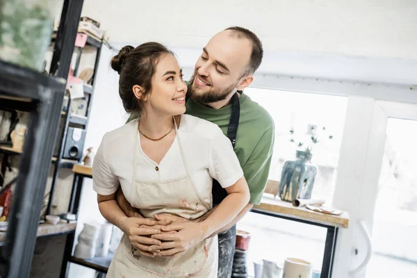 Smiling bearded artisan in apron hugging and looking at girlfriend in ceramic workshop — Stock Photo