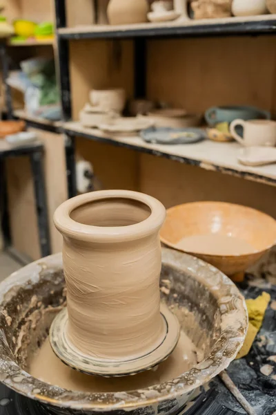Clay vase on pottery wheel near blurred rack in ceramic workshop at background — Stock Photo