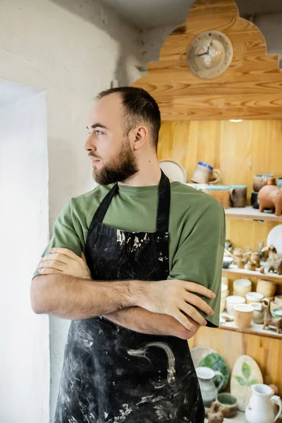 Artesano barbudo en delantal cruzando brazos y mirando hacia otro lado mientras está de pie en taller de cerámica - foto de stock